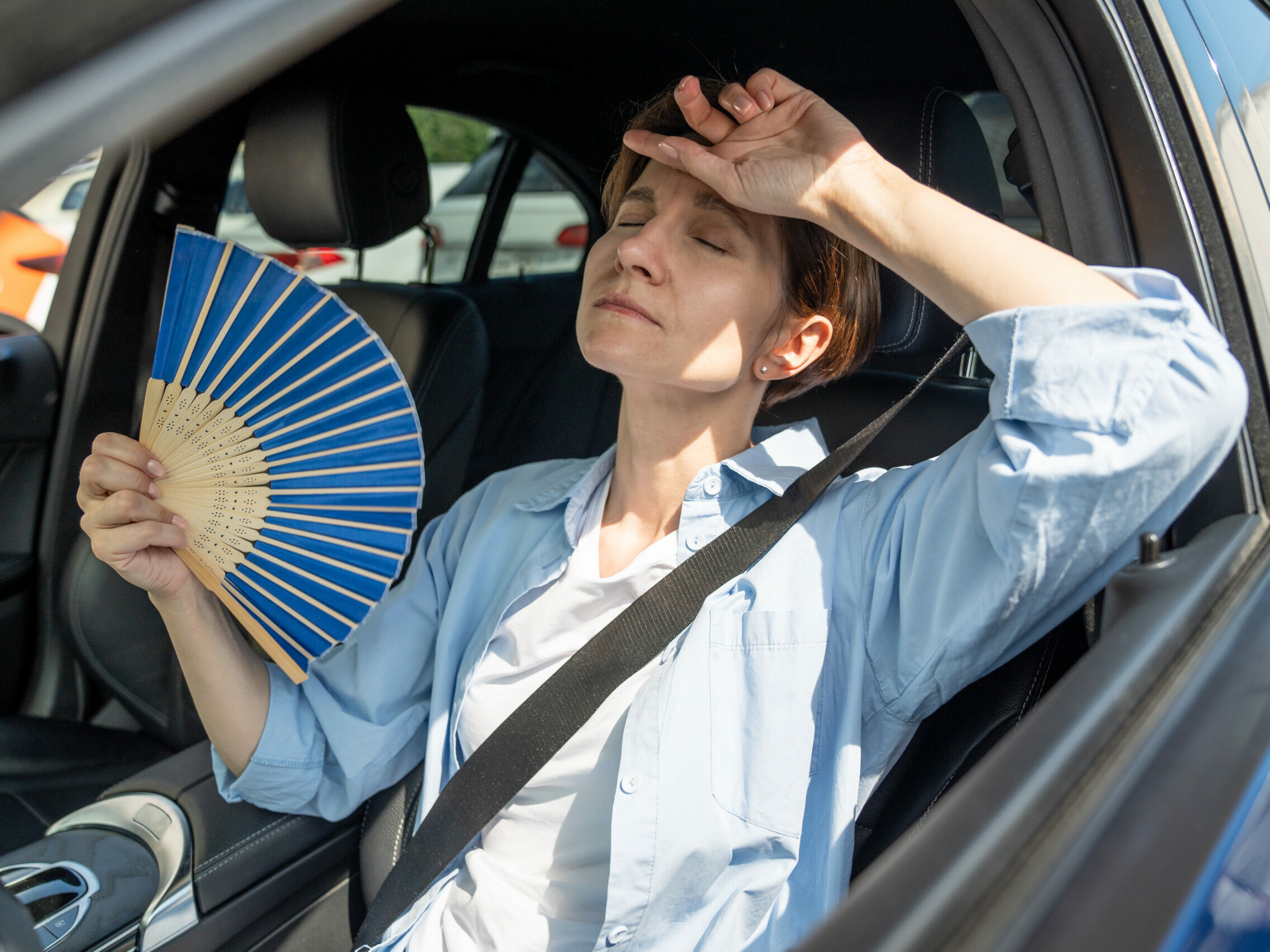 Exhausted woman sat in her car, with a paper fan in hand, suffering from extreme heat