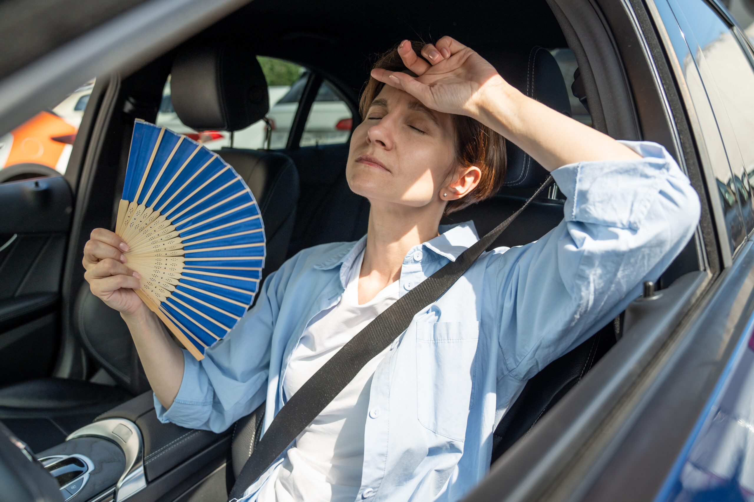 Exhausted woman sat in her car, with a paper fan in hand, suffering from extreme heat